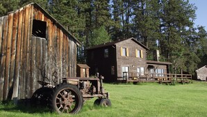 A rustic wooden barn and a two-story house with a porch, surrounded by a grassy field and pine trees. - Sky View Log Cabin- Off the beaten trail yet close to all the main attractions! (Custer)