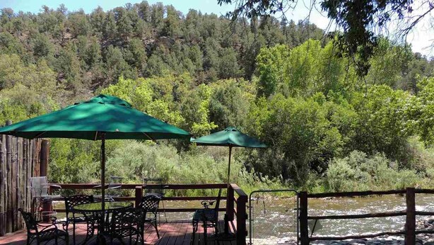 A wooden deck with outdoor furniture and umbrellas, overlooking a river and forested area. - On the Pecos River in Pecos New Mexico (Pecos)