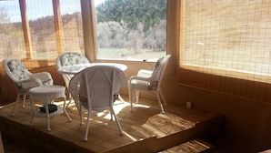 A sunlit room with wicker furniture, a small table, and a view of a forest through large windows. - On the Pecos River in Pecos New Mexico (Pecos)
