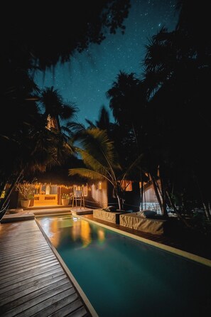 A nighttime pool area with a wooden deck, palm trees, and a thatched-roof structure.
