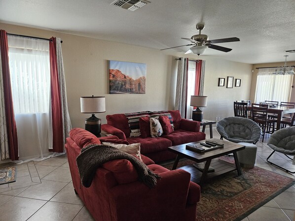 A living room with a red sofa, two lamps, a coffee table, a ceiling fan, and a dining area with chairs and a table.