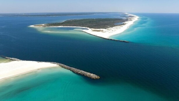 Luftaufnahme einer Küstenlandschaft mit sandigem Strand, klarem türkisblauem Wasser und einer kleinen Insel mit dichter Vegetation.