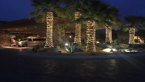 A building with a stone facade, palm trees wrapped in lights, and a parked vehicle in front.