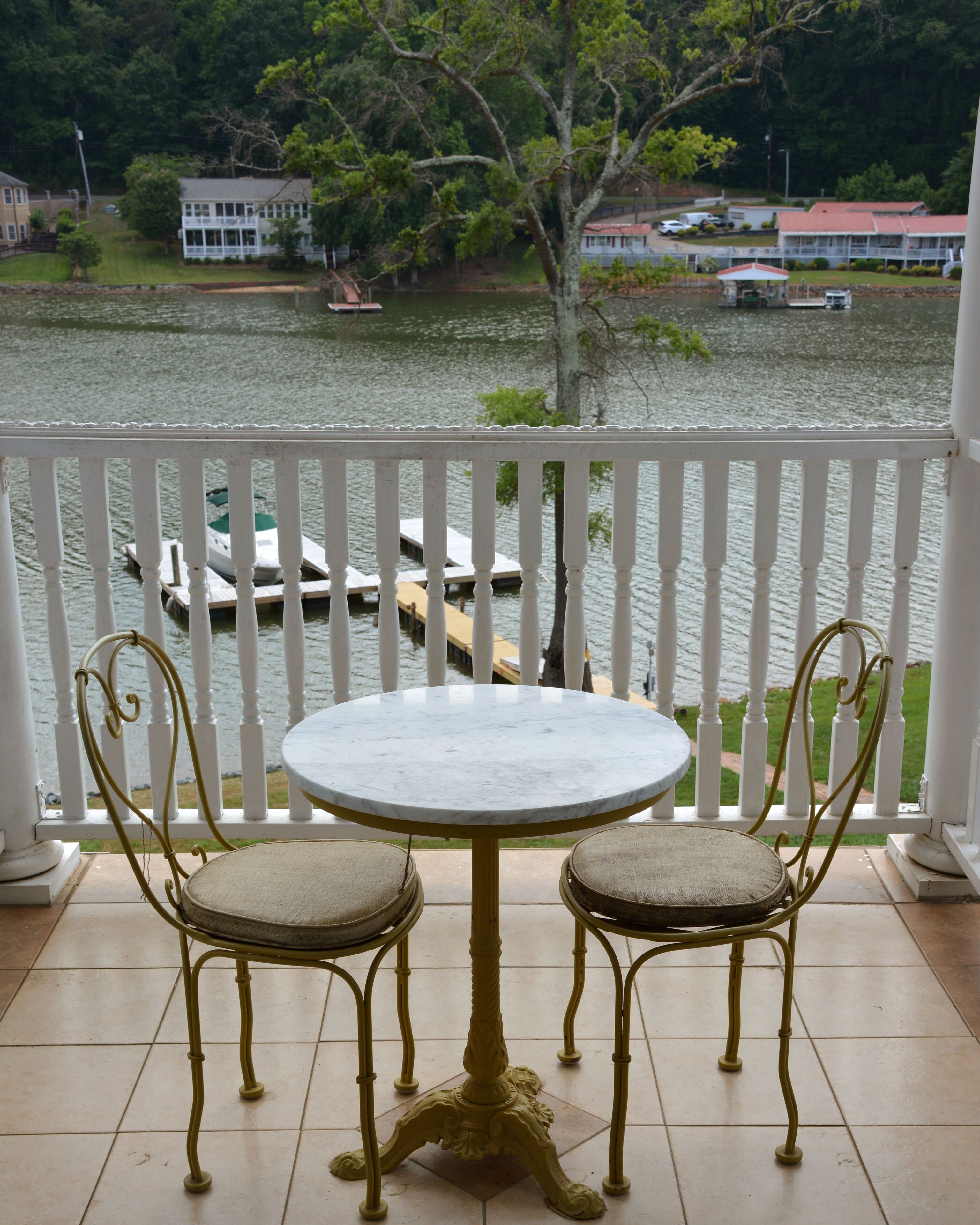 A balcony with a white railing, a round table with two chairs, and a view of a lake with boats and buildings in the background.