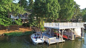 A dock with a boat and a wooden deck extending over water, surrounded by trees and houses. - Lakefront Luxe Retreat in Tranquil Cove  Endless Enjoyment  Lakeside Fireplace (New London)