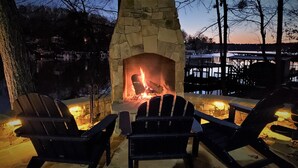A stone fireplace with a fire, surrounded by Adirondack chairs. - Lakefront Luxe Retreat in Tranquil Cove  Endless Enjoyment  Lakeside Fireplace (New London)