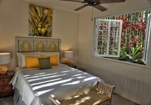 Une chambre à coucher comprenant un lit, deux tables de chevet, une commode, une chaise, un ventilateur de plafond et une fenêtre donnant sur des plantes.