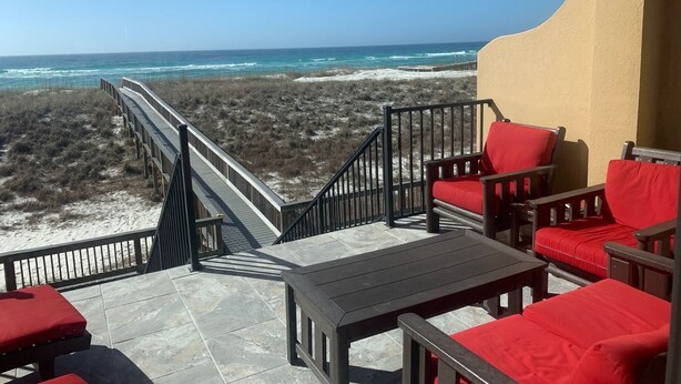 Un balcón con sillas rojas acolchadas y una mesa, con vistas a una playa que cuenta con un paseo marítimo de madera y dunas de arena.