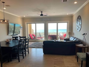 A spacious living area with a large sofa, a dining table with chairs, and a television. There is a balcony with red chairs and a view of the ocean.