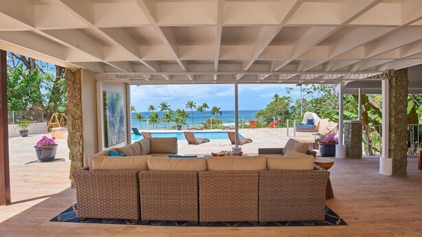 A covered outdoor seating area with a view of the ocean and palm trees.