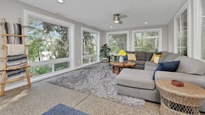 A spacious living room with large windows, a ceiling fan, a grey sofa, a coffee table, and a wicker side table.