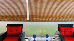 Un balcón con mobiliario de mimbre, dos cojines rojos y una mesa con dos vasos azules.