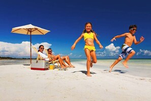 Eine Strandansicht mit Menschen, die unter einem Sonnenschirm entspannen, einem Kühlbox und einem Eimer.