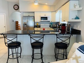 A modern kitchen with white cabinets, a stainless steel refrigerator, a microwave, and a coffee machine. There are three black bar stools and a white countertop. - HAPPYOURS Luxury Heated Pool, Hot tub No Additional Fees!!! @ Vanderbilt beach (Naples)