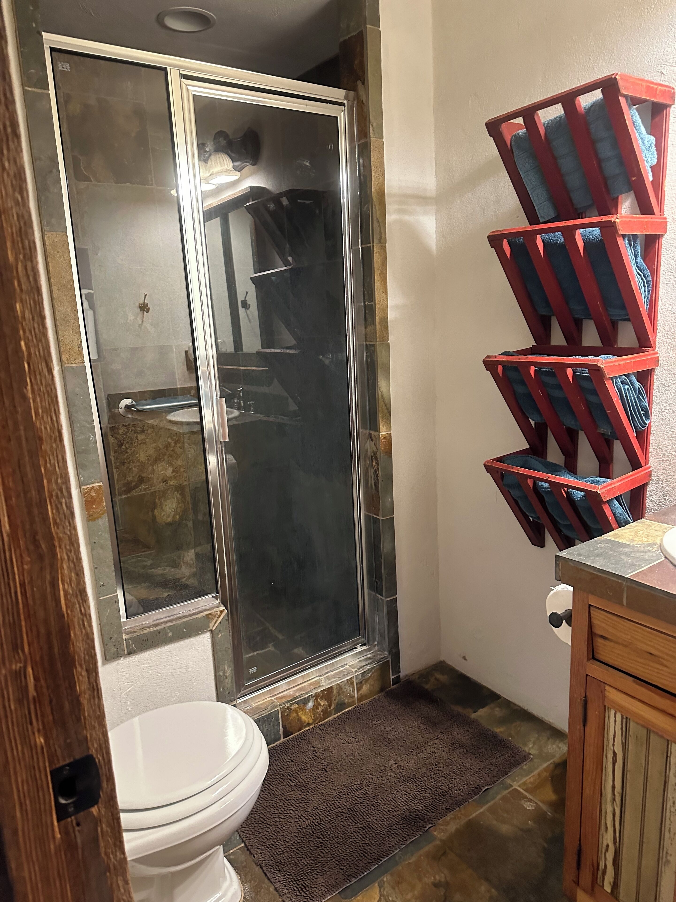 A bathroom with a glass shower enclosure, a white toilet, a wooden vanity, and a towel rack with multiple towels.