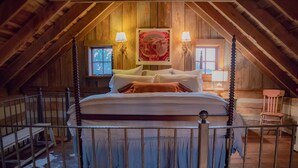 Une chambre mansardée avec un plafond en bois, un lit recouvert de draps blancs, deux lampes fixées au mur et un grand tableau encadré au-dessus du lit.