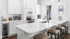 A modern kitchen with white cabinetry, a central island with stools, a built-in oven, and a large window. - Sunsets Look Better From Above at The Upper Deck / Lagoon Pool in Seacrest Beach / 4 Bikes (Rosemary Bch)