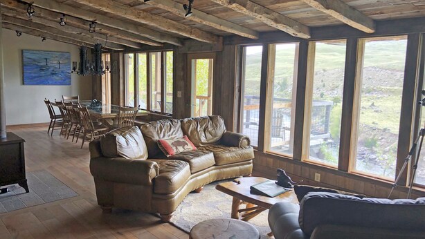 Een ruime woonkamer met een grote leren bank, een houten salontafel en een eethoek met een houten tafel en stoelen. Grote ramen bieden uitzicht op het omliggende landschap.
