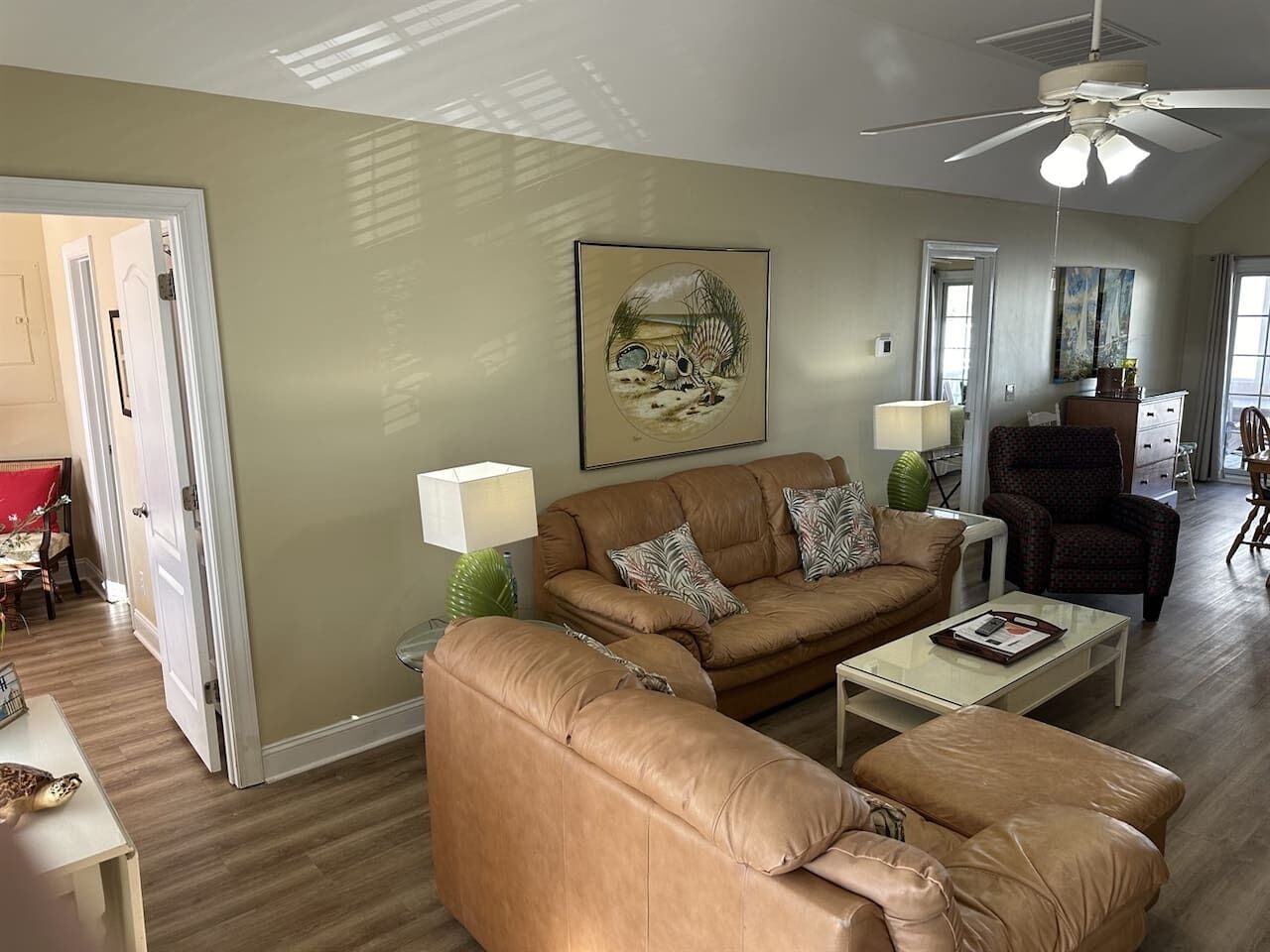 Un salon avec un canapé en cuir marron, une table basse et un ventilateur de plafond. Il y a un tableau au mur et une porte qui donne sur une autre pièce.