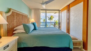 A hotel room with a bed, a ceiling fan, a nightstand, a lamp, a window with a view of palm trees, and a balcony. - Beachfront Mana Kai Maui Unit #502 (Kihei)