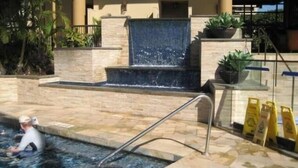 A swimming pool with a waterfall feature, surrounded by stone steps and potted plants. - Beachfront Mana Kai Maui Unit #502 (Kihei)