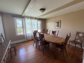 Une salle à manger avec une table et des chaises en bois, de grandes fenêtres avec des rideaux et un luminaire au plafond.