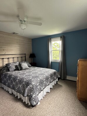 Ein Schlafzimmer mit einem Bett, einem Deckenventilator, einem Fenster mit Vorhängen und einem Nachttisch mit Lampe.