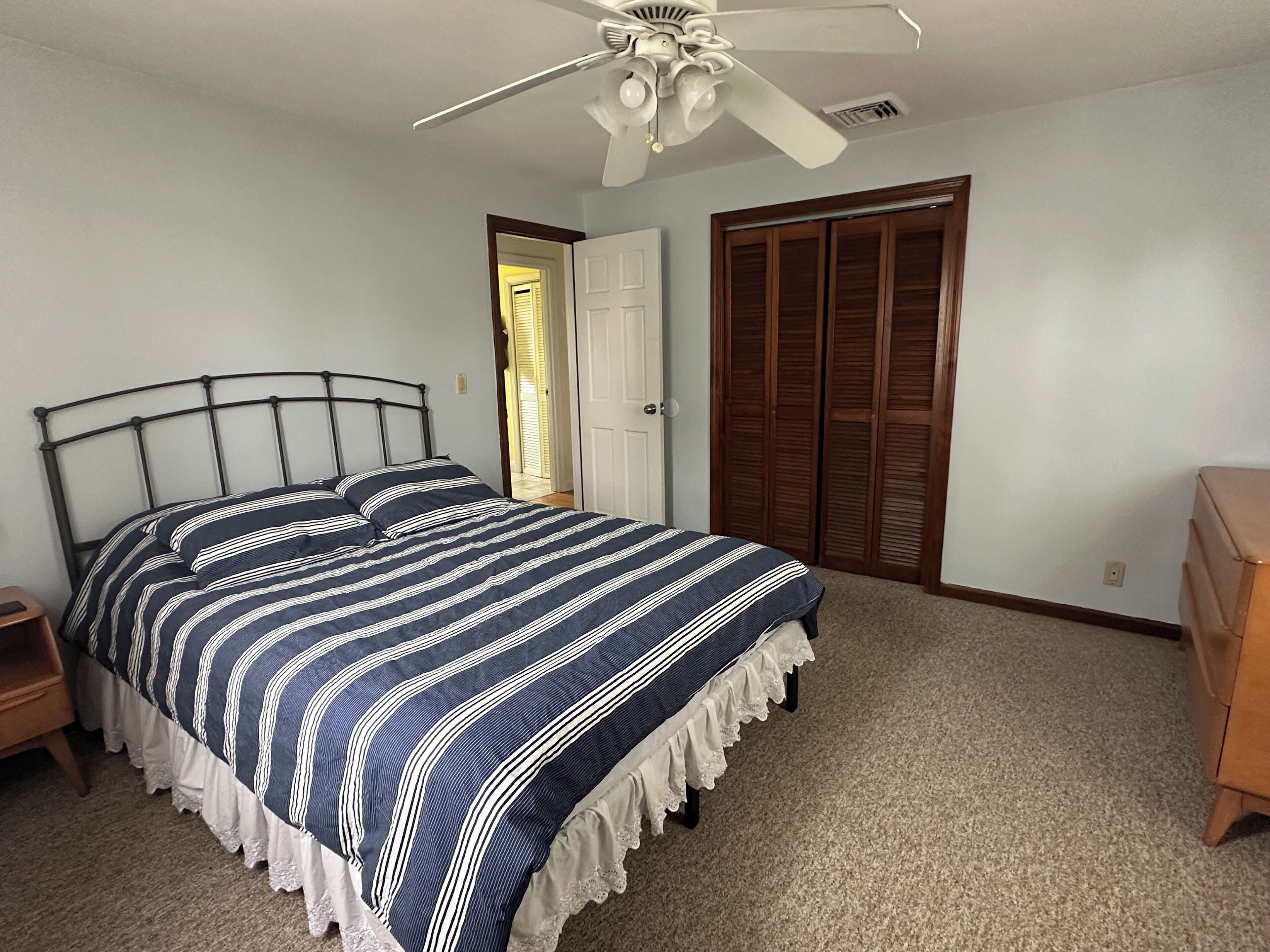Een slaapkamer met een bed, nachtkastje, dressoir, plafondventilator en een deur naar een andere kamer.