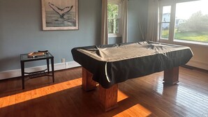 Una mesa de billar en una habitación con un ventanal grande, una obra de arte enmarcada en la pared y una pequeña mesa con una lámpara.