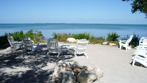 A beach with white Adirondack chairs arranged in a semi-circle around a fire pit. -  Spring & summer dates available licensed rental vr #25-0235  NO BOATS (tavernier)