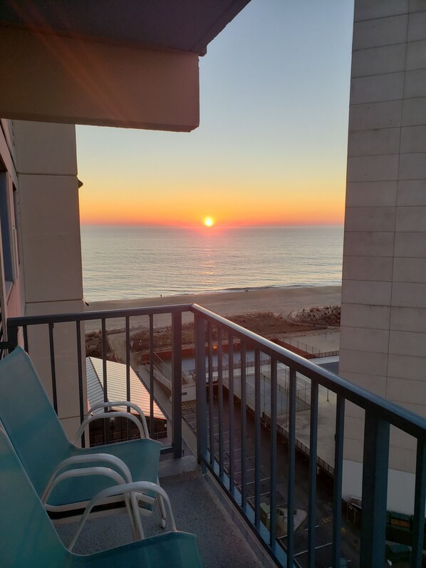A balcony with a view of the sunset over the sea, a railing, and two blue chairs.
