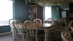 A dining area with a wooden table and white chairs, a bookshelf, and a view through a window.
