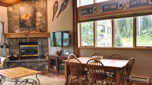 A stone fireplace with a TV, wooden dining table and chairs, and a large window with a view of trees. - Ski In/Ski out - Great Location in Big Sky Mountain Village (Big Sky)