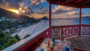 Une terrasse en bois avec une table dressée pour le dîner, offrant une vue sur une plage et un paysage montagneux au coucher du soleil.