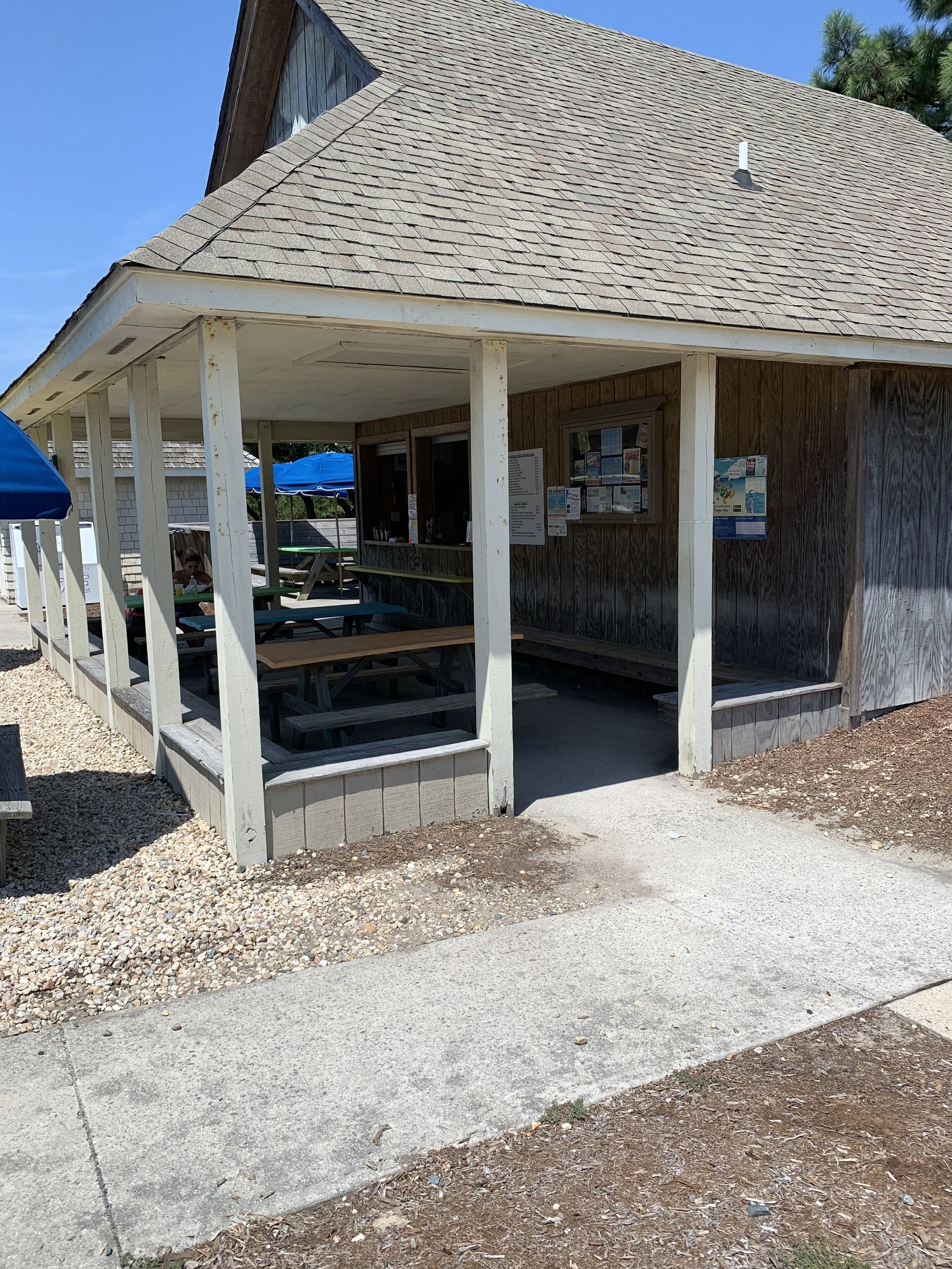 A covered picnic area with tables and benches, a wooden building with a sloped roof, and a gravel ground.
