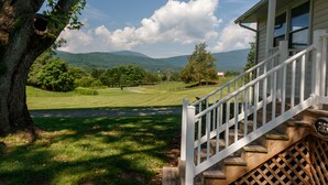 A house with a wooden staircase leading to a porch, a large tree with a hollow trunk, and a view of a green field and mountains in the background. - Relax in the beautiful South River Valley just minutes from wineries,  (Stanardsville)
