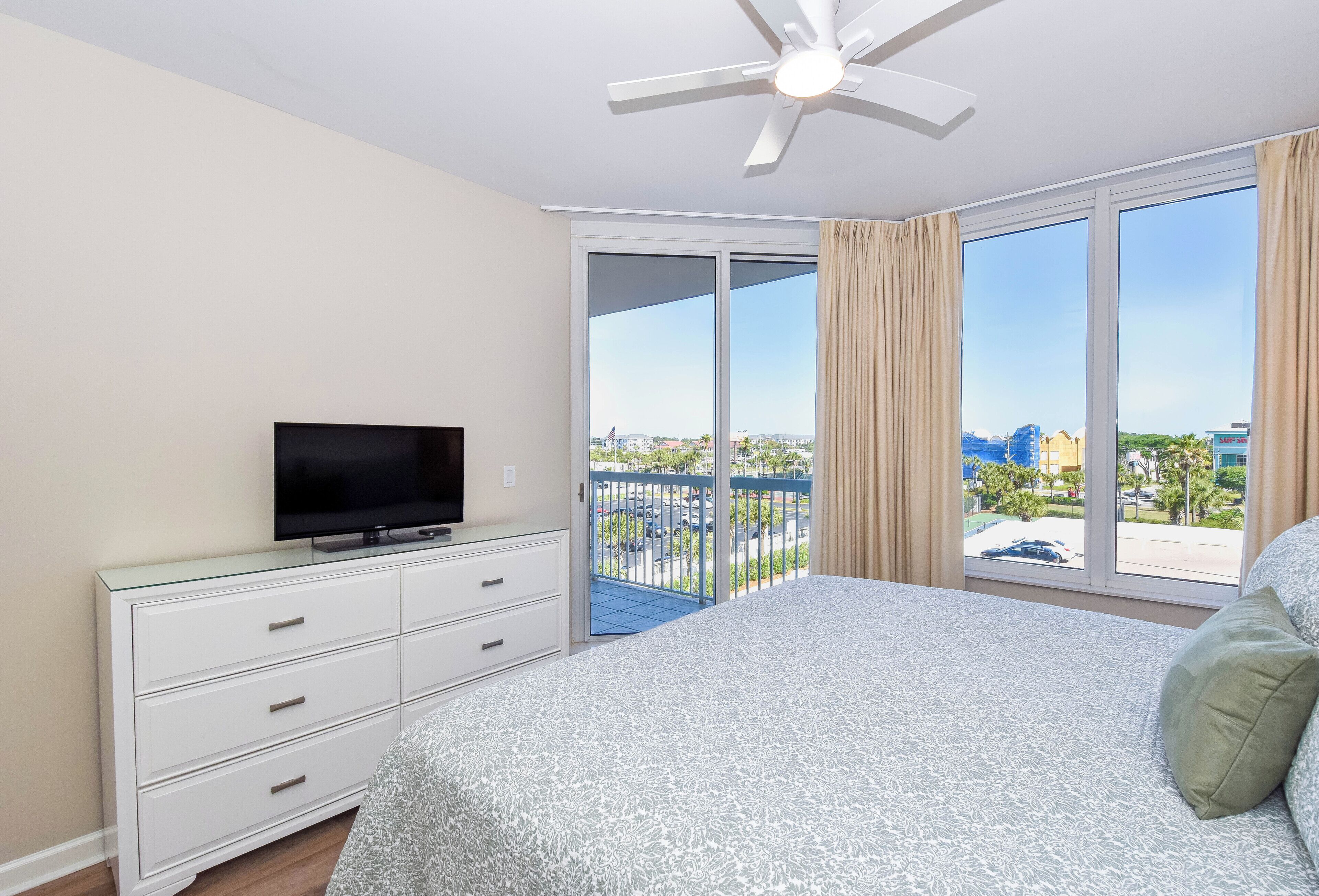 A bedroom with a bed, a TV on a dresser, a balcony with a view, and a ceiling fan.