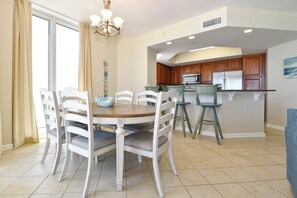 Un comedor con una mesa redonda y sillas blancas, una cocina con armarios de madera y un refrigerador, y un ventanal con cortinas.