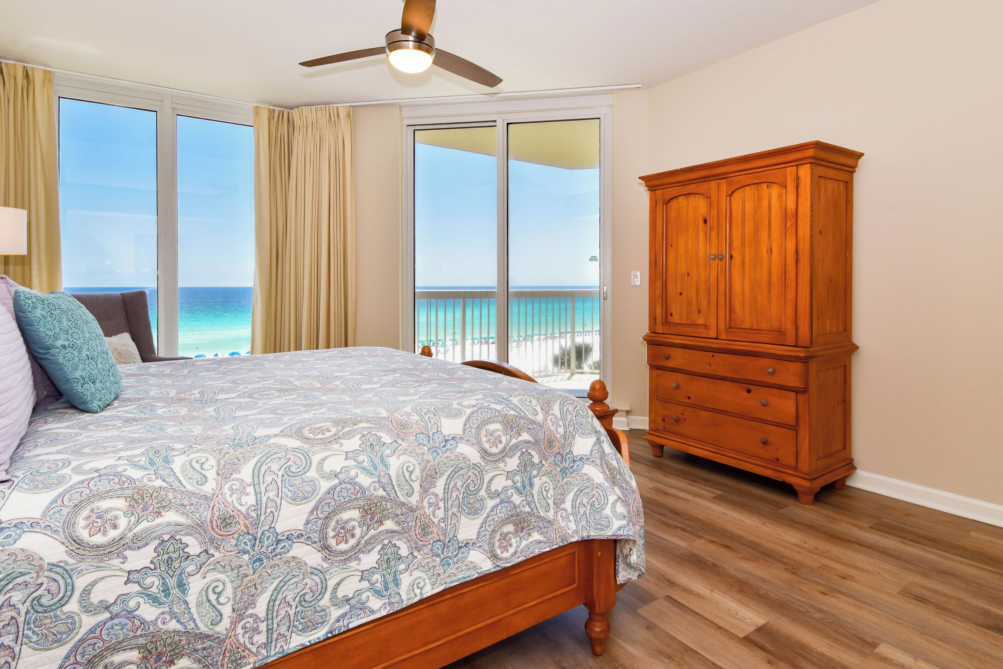 A bedroom with a large bed, a wooden cabinet, and a view of the ocean through a sliding glass door.