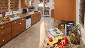 Una cucina con piani di lavoro in granito, mobili in legno, pavimento in pietra e vista sull'oceano tramite una porta scorrevole in vetro.