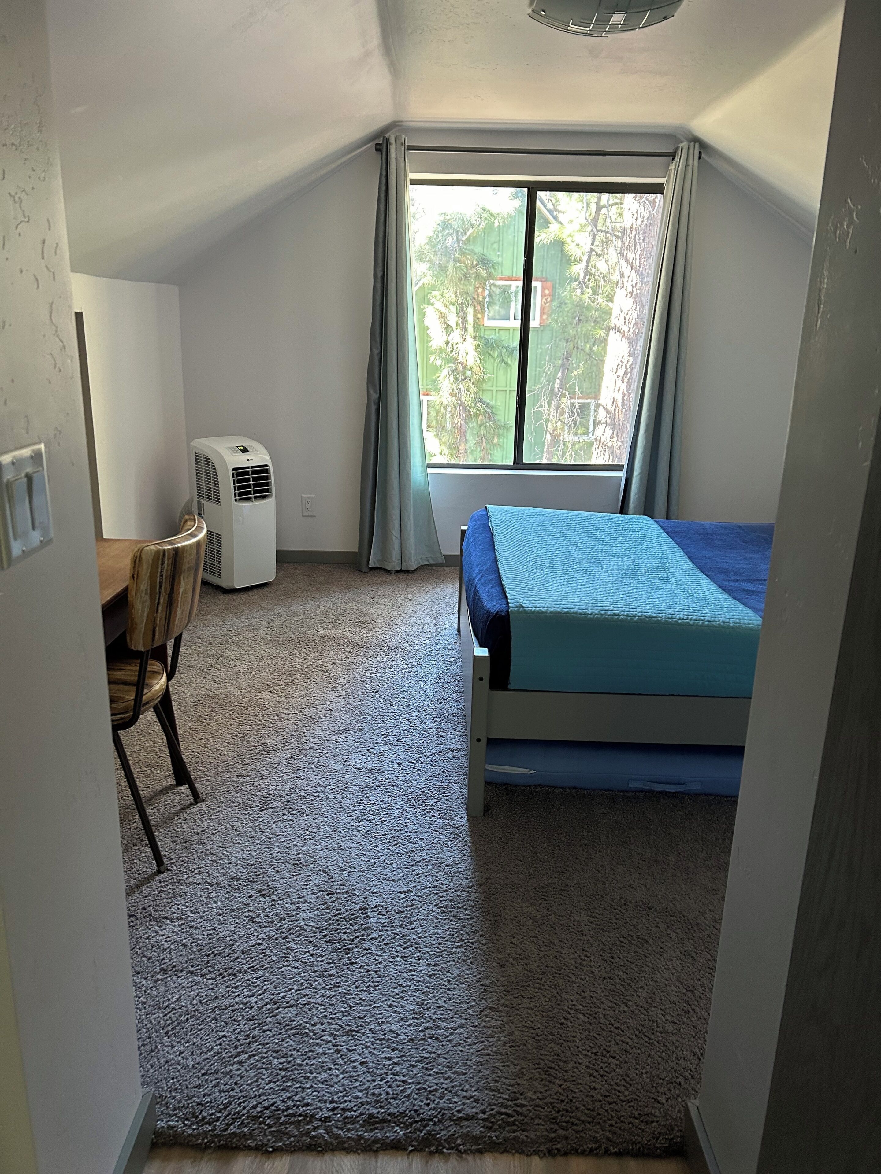Una habitación pequeña y bien iluminada con una cama individual, un escritorio con una silla y una ventana con cortinas.