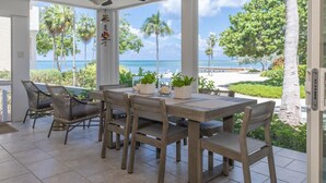 Zona pranzo con vista sulla spiaggia e sull'oceano, con tavolo e sedie, ventilatore a soffitto e piante in vaso.