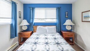 Un dormitorio con una cama, dos mesitas de noche con lámparas, una ventana con cortinas azules y una pared con un cuadro enmarcado.