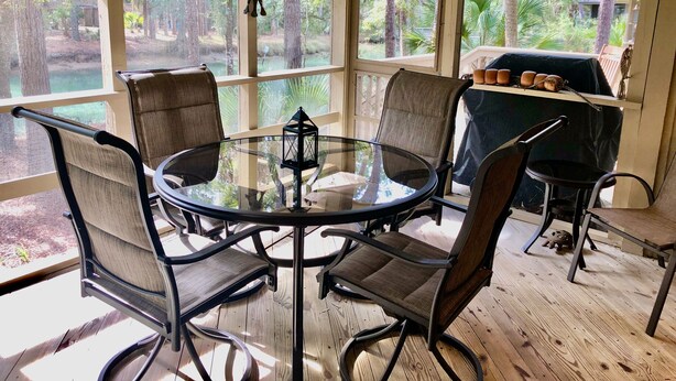 A sunlit room with a round glass table, four chairs, and a view of a forest through large windows. - Inlet Cove Paradise #36 (Kiawah Island)