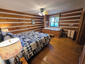 A cozy cabin bedroom with a wooden floor, a bed with a patterned quilt, a bedside lamp, a window with curtains, and a ceiling fan. - Maggie’s House Cabin: A Cozy Retreat with Mountain Views, Hot Tub, and Jacuzzi (Sieverville)