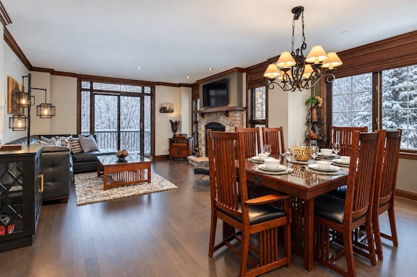 A dining area with a wooden table set for six, flanked by wooden chairs, a fireplace, and a television.