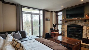 A bedroom with a large bed, a fireplace, a television, and a view of trees outside.