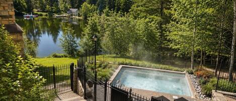 Una piscina rectangular con borde de piedra, rodeada por una cerca de metal negro, y vista a un lago con árboles y una casa al fondo.