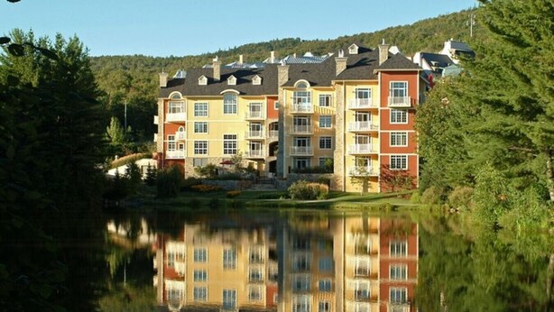 A multi-story hotel with balconies, a reflection in the water, and a forested hillside in the background.
