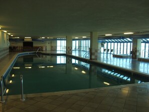 An indoor swimming pool with clear glass walls, tiled floor, and a few chairs around the pool area.